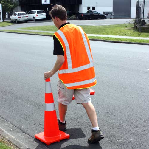 Aerocone Orange 900mm, a slim profile safety cone, is displayed on the street. A man wearing a reflective vest and shorts walks nearby, emphasizing the cone's stability and visibility.