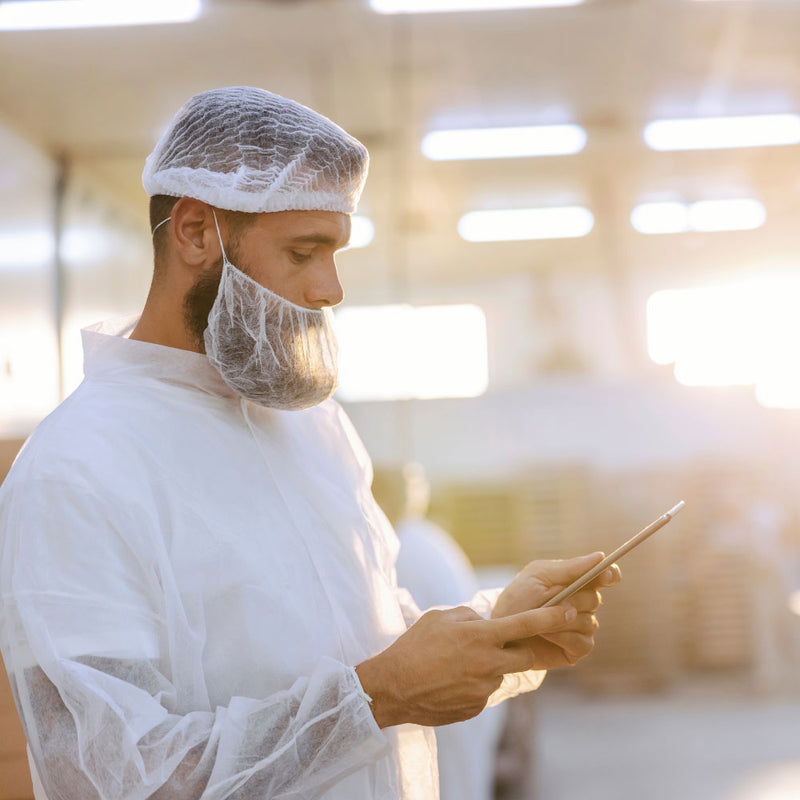 Load image into Gallery viewer, Worker in hygiene gear reviews data on tablet in a processing facility, showcasing Bastion Disposable Beard Protector, Single Loop: Carton/1000, emphasizing hygiene and secure fit in industrial environments.