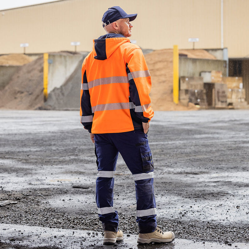 Load image into Gallery viewer, Man in industrial yard wearing Bisley Flex & Move Hi Vis Taped Water Repellent Hoodie, featuring reflective stripes and bio-motion tape, paired with dark blue cargo trousers and beige work boots.