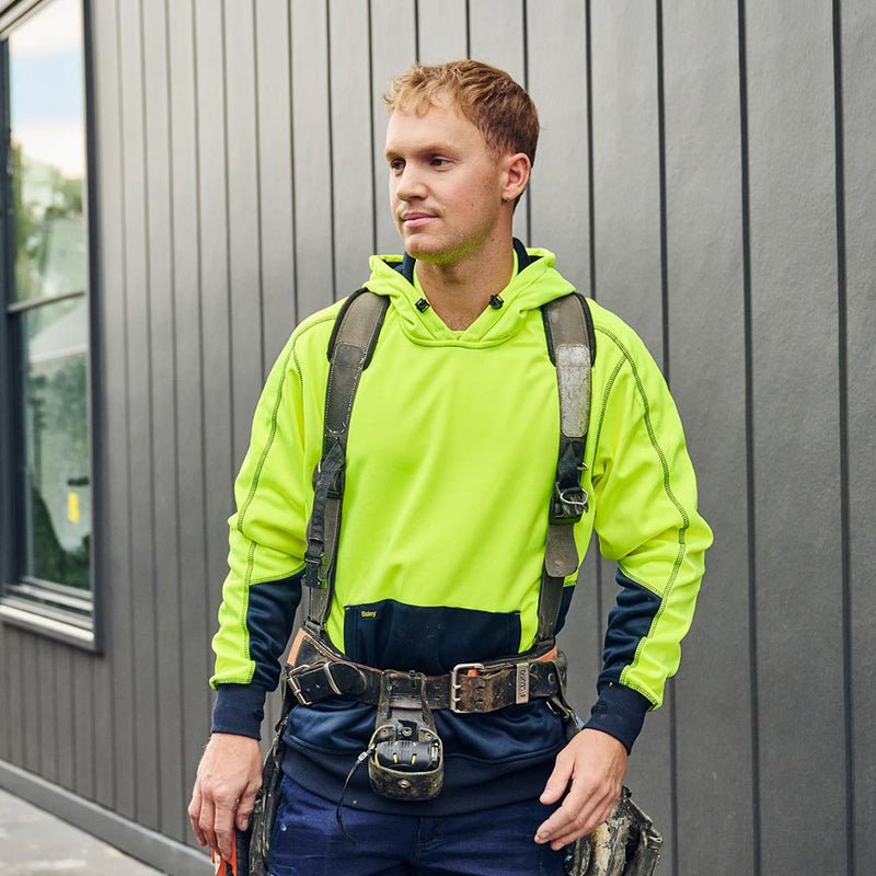 Load image into Gallery viewer, A man in a Bisley Hi Vis Pullover Fleece Hoodie stands beside a building, showcasing the hoodie’s shaped hood, kangaroo pouch pocket, and safety harness, reflecting Active Safety's commitment to reliable workwear.