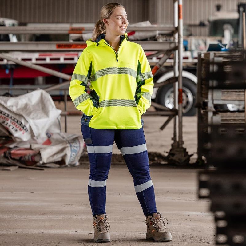 Load image into Gallery viewer, A woman in a workshop wears the Bisley Women's Flex & Move Hi Vis Taped Water Repellent Hoodie, featuring reflective bands, a shaped hood, and zipped pockets, ideal for safety and comfort.