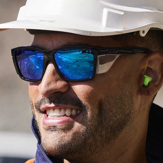 A man smiling while wearing personal protective safety equipment. Wearing a white hard hat on his head, Bolle Safety Glasses to protect his eyes and earplugs for hearing protection.