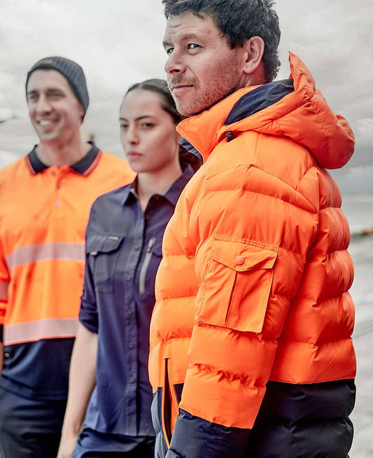 Three people wearing hi vis work uniforms. One is wearing a hi vis orange polo shirt, another is wearing a navy work shirt and the other is wearing a hi vis puffer jacket to keep warm in the workplace.