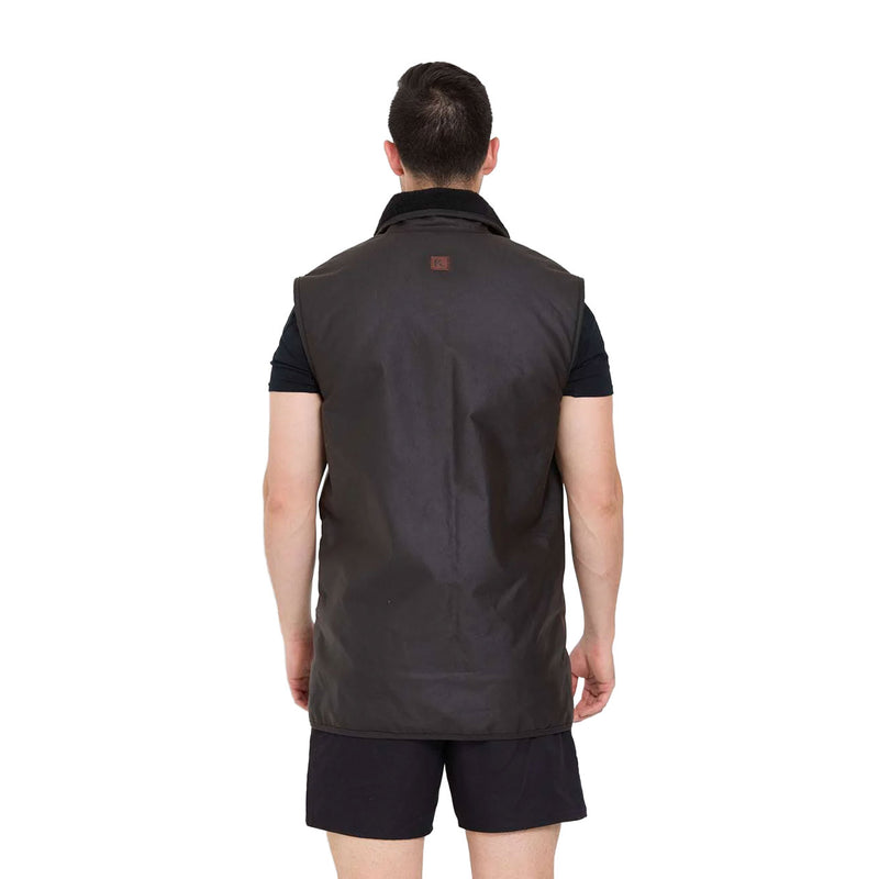Load image into Gallery viewer, Back view of a man wearing the Kaiwaka Sleeveless Oilskin Vest over a black shirt and shorts, showcasing its rugged design and functionality for outdoor work, emphasizing mobility and durability.
