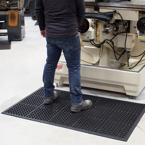 Safety Cushion Mat: 1500 x 900mm, shown with a person standing on it, is a heavy-duty, non-skid anti-fatigue mat with beveled edges and holes for spill drainage, ideal for industrial environments.