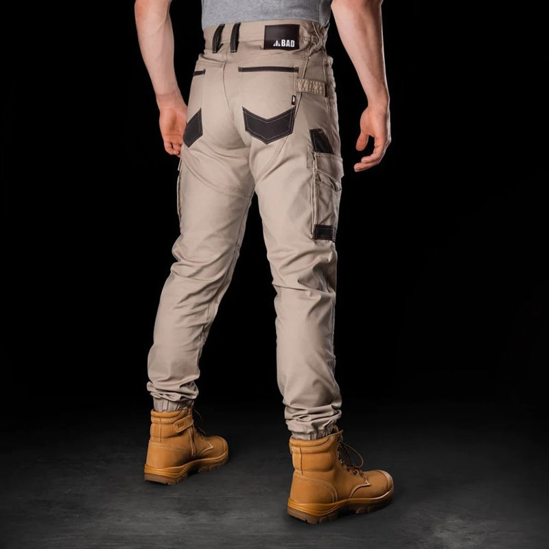 Load image into Gallery viewer, BAD Attitude Slim Fit Cuffed Work Pants, featuring reinforced pockets and knees, are worn by a man standing in brown boots, showcasing the pants' utilitarian design suitable for active work environments.
