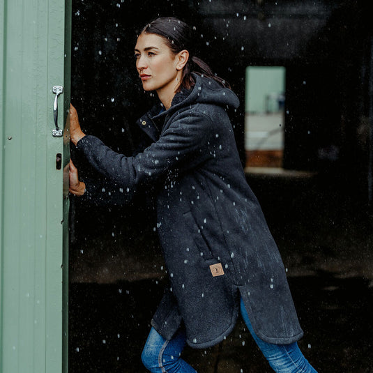 Hard Yakka Womens Longline Zip Hoodie, Black, worn by a woman stepping through a door, showcasing its longline design, curved hemline, and practical pockets, ideal for winter warmth and workplace comfort.
