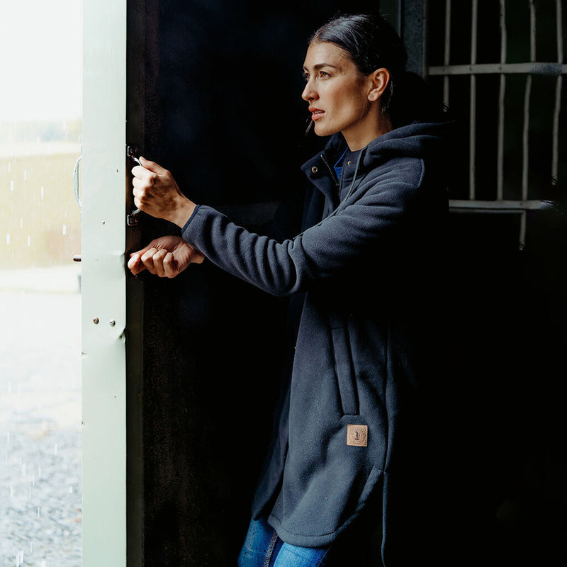 Load image into Gallery viewer, Hard Yakka Womens Longline Zip Hoodie, Black, worn by a woman opening a door, featuring a longline design with curved hem, side splits, and a hood, emphasizing warmth and comfort.