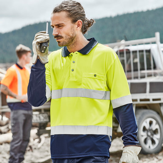 Syzmik Hi Vis Taped L/S Spliced Polo Shirt: A man in a yellow, long-sleeved high-visibility shirt with reflective tape, holding a walkie-talkie, demonstrating durable, moisture-wicking workwear ideal for safety-focused environments.