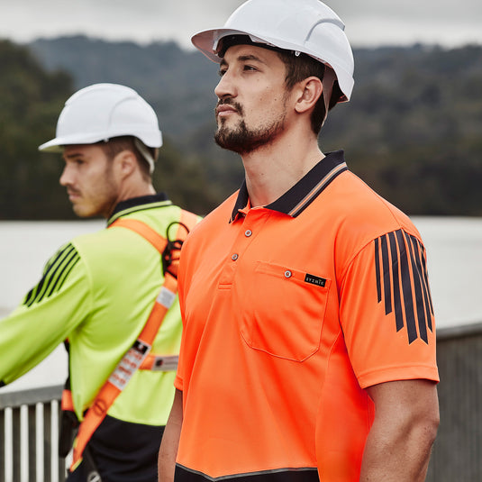 Syzmik Flux Hi Vis Polo Shirt worn by men in hard hats, features breathable mesh panels for hot conditions, chest pocket with pen insert, showcasing versatility for safety-focused work environments.