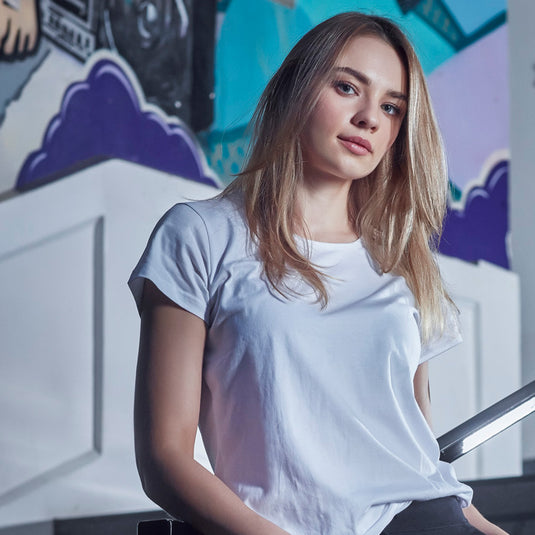 Syzmik Womens Streetworx T-Shirt showcased on a model, highlighting its modern slim fit and scoop neck design, perfect for professional workwear environments. The shirt offers a longer tail with a curved hem.