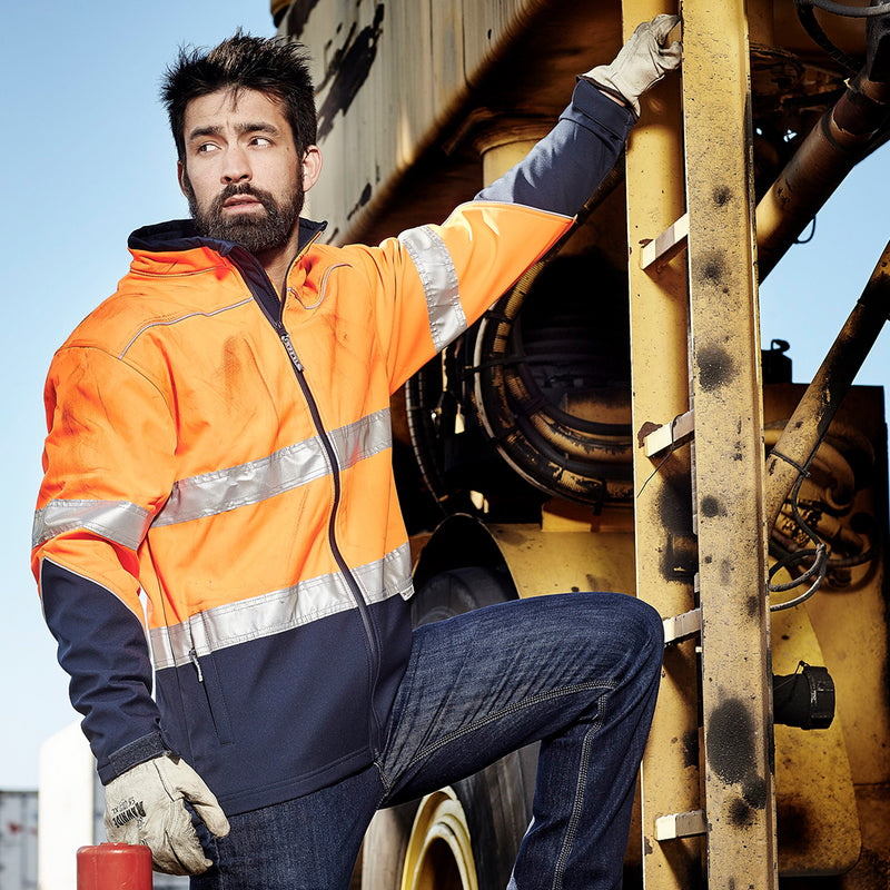 Load image into Gallery viewer, Syzmik Hi Vis Taped Soft Shell Jacket showcased on a man leaning on a yellow structure, highlighting its waterproof zips, reflective tape, and adjustable cuffs, ideal for safety and workwear environments.