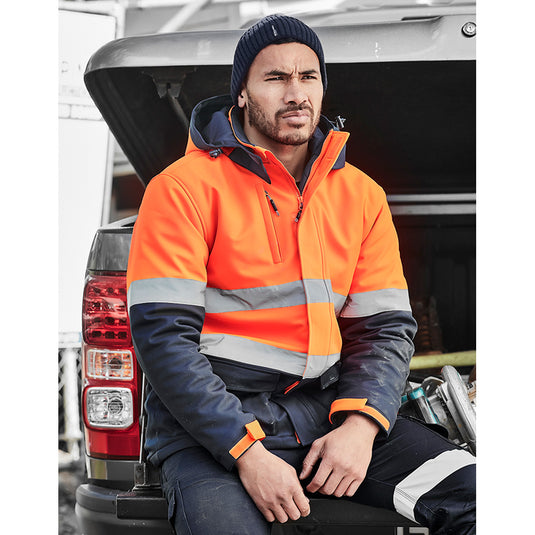 Syzmik Hi Vis Antarctic Softshell Taped Jacket worn by a man in a truck, showcasing its longer length and functional design with visible pockets and hood, ideal for cold conditions and safety.
