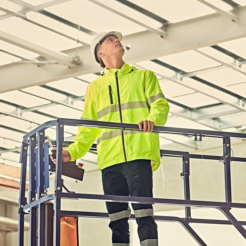 Load image into Gallery viewer, A worker on a scissor lift inspects overhead beams in an industrial building, wearing a Syzmik Unisex Super Light Segmented Tape Packable Rain Jacket, showcasing its practicality and safety features for active environments.