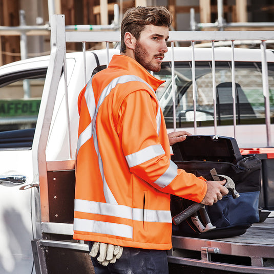 Syzmik 2-in-1 X Back Soft Shell Jacket worn by a man, featuring a tool bag, detachable sleeves, and twin hoop bio motion tape for enhanced visibility, ideal for construction settings.