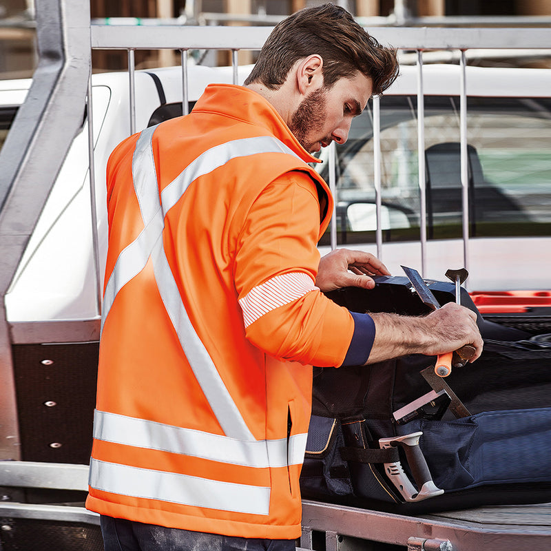 Load image into Gallery viewer, Syzmik 2-in-1 X Back Soft Shell Jacket, worn by a man examining a tool, features detachable sleeves, high-visibility tape, and multiple pockets, ideal for safety and functionality.