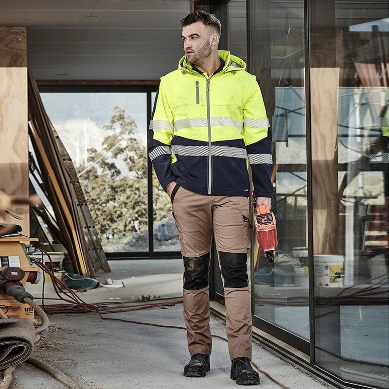 Load image into Gallery viewer, Syzmik Streetworx Tough Pant showcased on a man holding a power saw, featuring stretch fabric, durable knee reinforcement, and multiple functional pockets, ideal for safety-focused work environments.