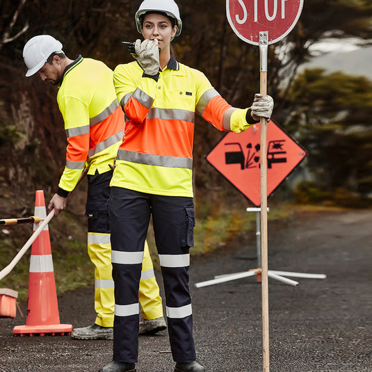 Syzmik Womens Bio Motion Taped Pant, showcased by a woman in safety gear holding a stop sign, features cargo and mobile pockets, tailored fit, and reflective tape for visibility. Ideal for active safety environments.