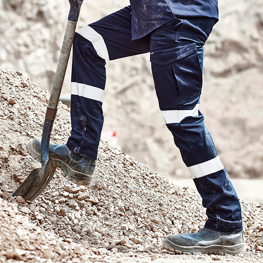 Syzmik Essential Stretch Taped Cargo Pant worn by a person digging, showcasing its durability and functionality with reflective tape, ample pockets, and comfortable slim fit. Ideal for outdoor work environments.