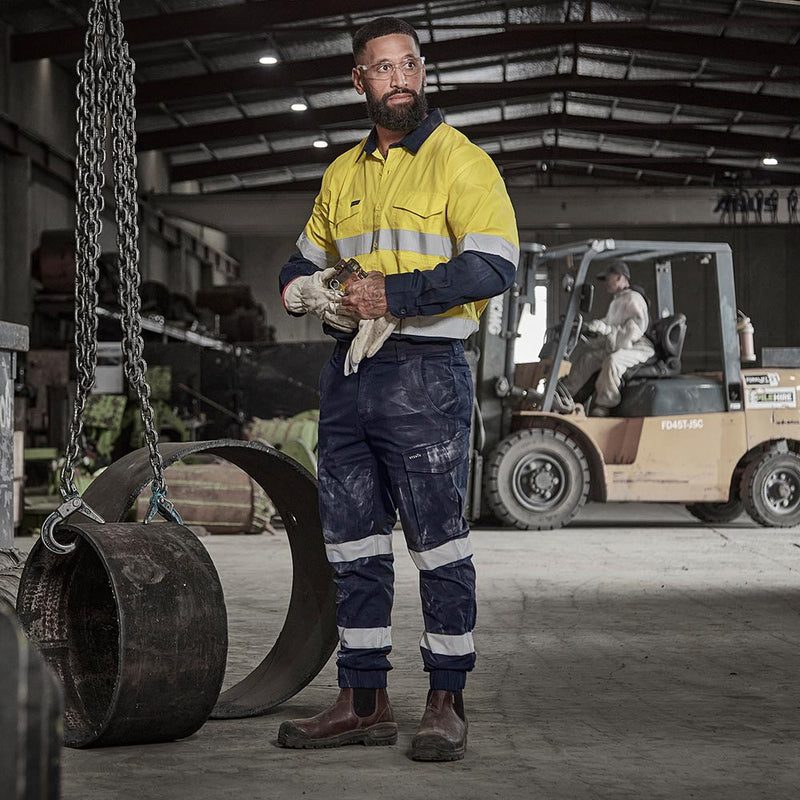 Load image into Gallery viewer, Syzmik Essential Stretch Taped Cuffed Cargo Pant, Navy, showcased on a worker in a warehouse. Features include reflective stripes, triple stitching, multiple pockets, and elastic waistband for comfort and safety.