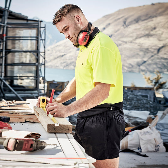 Man in a yellow shirt measures wood while wearing Syzmik Cotton Rugby Shorts, showcasing their lightweight cotton twill fabric and practical design for comfort and utility in construction settings.