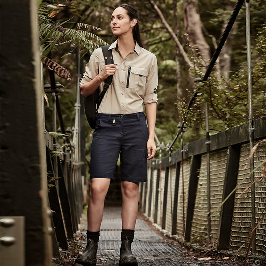 Syzmik Womens Rugged Cooling Vented Short, showcased on a woman walking on a bridge, features multi-functional pockets and reinforced stress points, ideal for active work environments in safety and workwear settings.