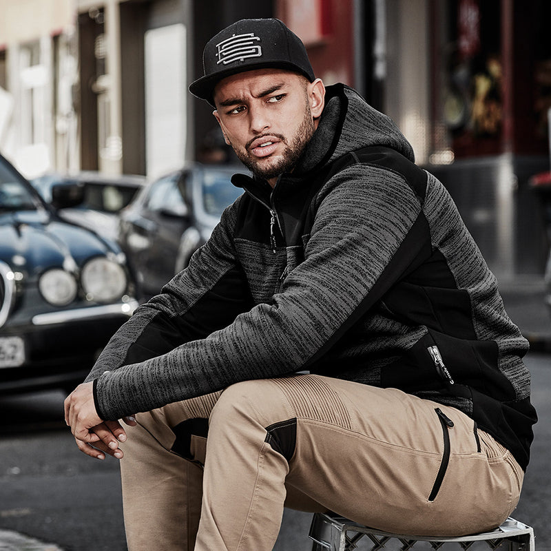 Load image into Gallery viewer, Syzmik Streetworx Knit Hoodie worn by a man sitting, showcasing reinforced soft shell panels and built-in thumb holes, ideal for workplace safety and comfort.
