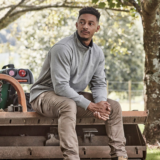 Syzmik 1/4 Zip Brushed Fleece Jumper shown worn by a man seated on a bench, highlighting its practical half-zip design and zippered pockets, ideal for secure storage in active environments.