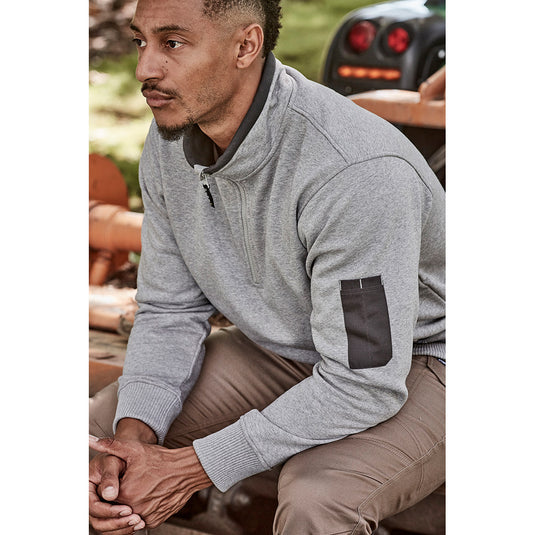 Syzmik 1/4 Zip Brushed Fleece Jumper on a man seated on a truck, showcasing its soft fleece texture, half-zip design, and zippered hip pockets for secure storage, ideal for cold outdoor workdays.