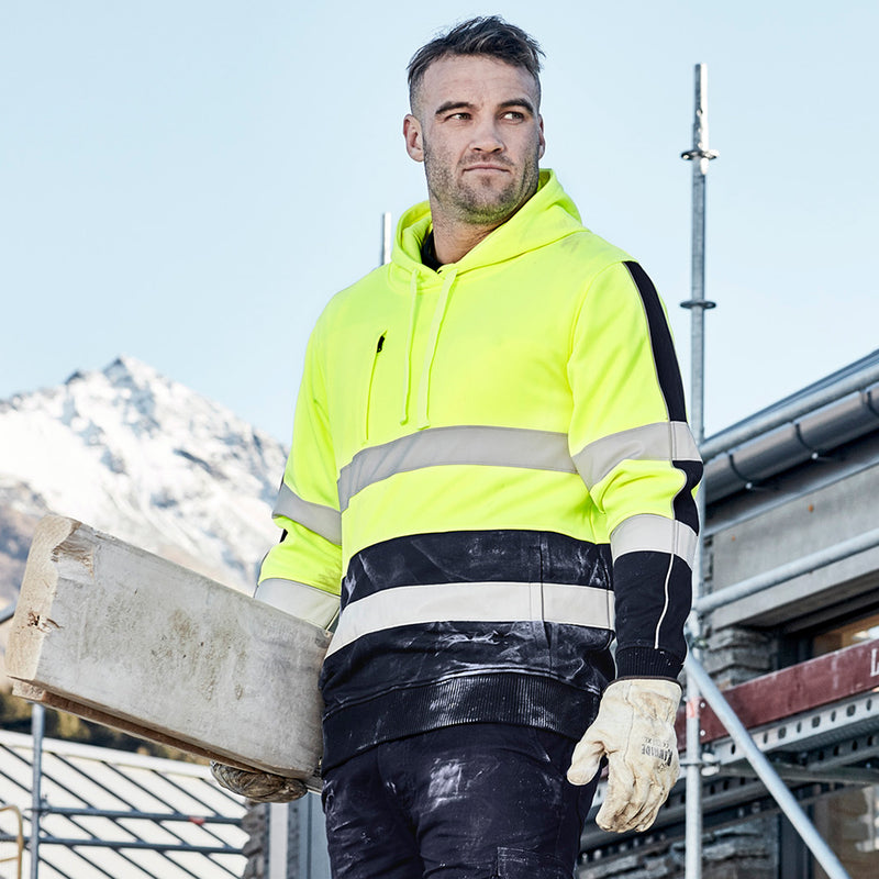 Load image into Gallery viewer, Syzmik Hi Vis Stretch Taped Hoodie worn by a man outdoors, holding wood, showcasing its reflective tape and durable design, ideal for safety and work environments.