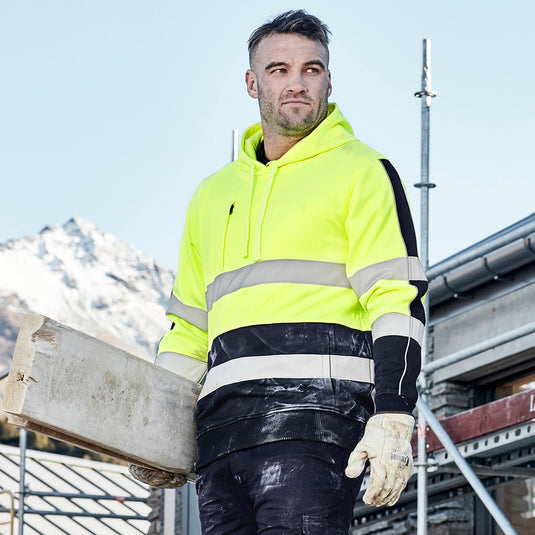 Syzmik Hi Vis Stretch Taped Hoodie worn by a man outdoors, holding wood, showcasing its reflective tape and durable design, ideal for safety and work environments.