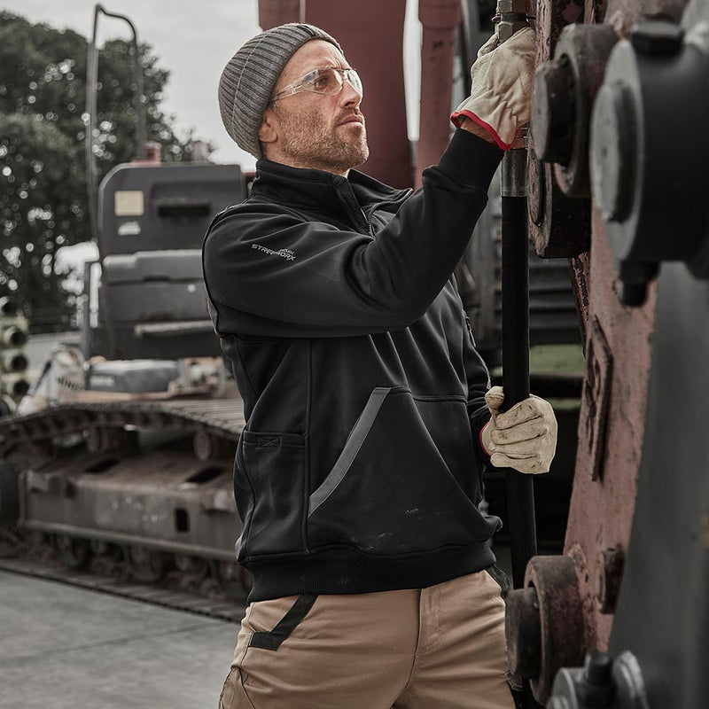 Load image into Gallery viewer, Man at industrial site wearing the Syzmik Streetworx Water Resistant 1/4 Zip Fleece Pullover, adjusting machinery. Features zippered kangaroo pockets, arm pocket, and eco-friendly water-resistant fabric, ideal for safety-focused work environments.