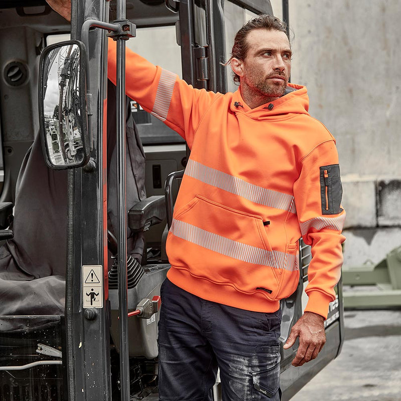 Load image into Gallery viewer, Syzmik Streetworx Hi Vis Water Resistant Taped Pullover Hoodie worn by a man standing in construction equipment doorway, showcasing reflective bands, zippered kangaroo pockets, and pencil pocket for workplace utility.