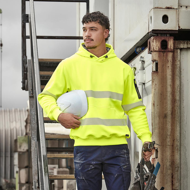 Load image into Gallery viewer, A man at a construction site wears the Syzmik Streetworx Hi Vis Water Resistant Taped Pullover Hoodie, featuring reflective stripes and carrying safety gear, highlighting its utility and protective qualities.