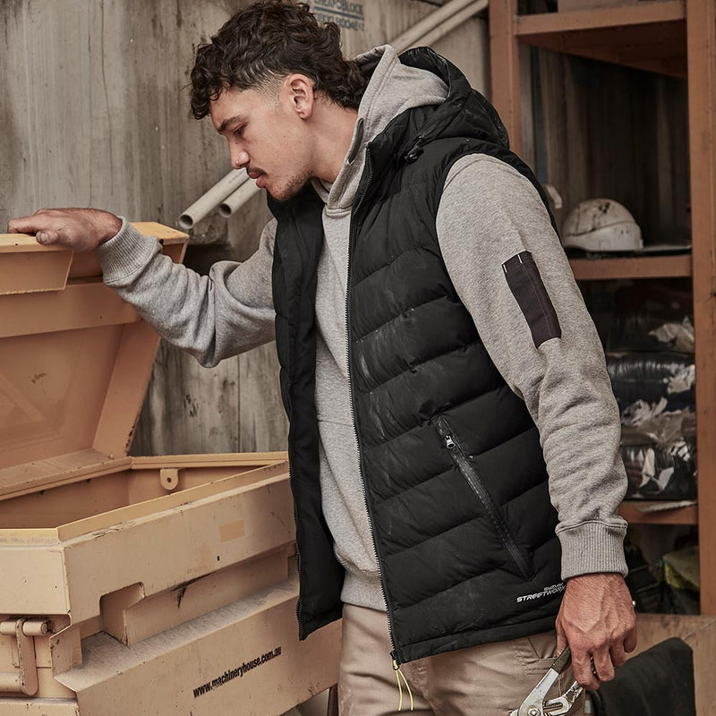 Load image into Gallery viewer, Young man in a work environment wearing the Syzmik Streetworx Hooded Puffer Vest, showcasing its durable quilted design and practical zip-off hood, while holding pliers next to an industrial storage bin.