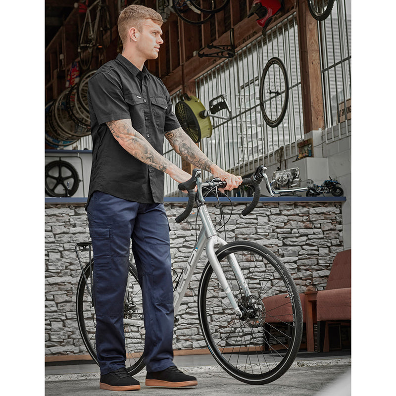 Load image into Gallery viewer, Syzmik Midweight Drill Cargo Pant, worn by a tattooed man holding a bicycle, showcasing its durability with multiple pockets, reinforced areas, and wide belt loops, ideal for safety and work environments.
