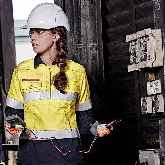 Syzmik Womens FR Closed Front Taped L/S Shirt worn by a woman in a hard hat, featuring gusset sleeves, chest pockets, and FR tape, ideal for construction and safety environments.
