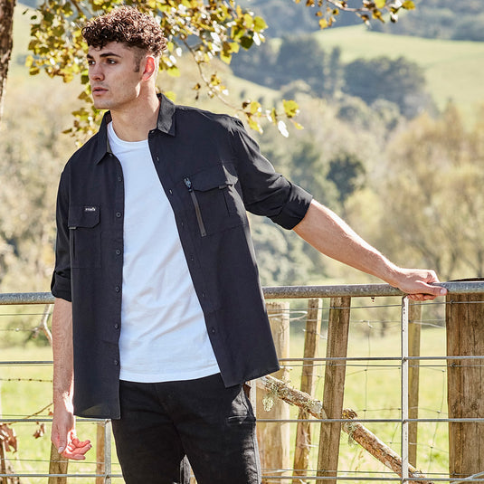Syzmik Outdoor Long Sleeve Work Shirt, worn by a man leaning on a fence, featuring roll-up tab sleeves, two chest pockets, and breathable fabric with underarm mesh for enhanced airflow.