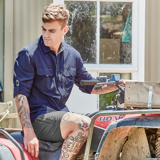 Syzmik Outdoor Long Sleeve Work Shirt worn by a tattooed man sitting on a motorcycle, illustrating its lightweight design with roll-up sleeves and chest pockets, ideal for active safety environments.