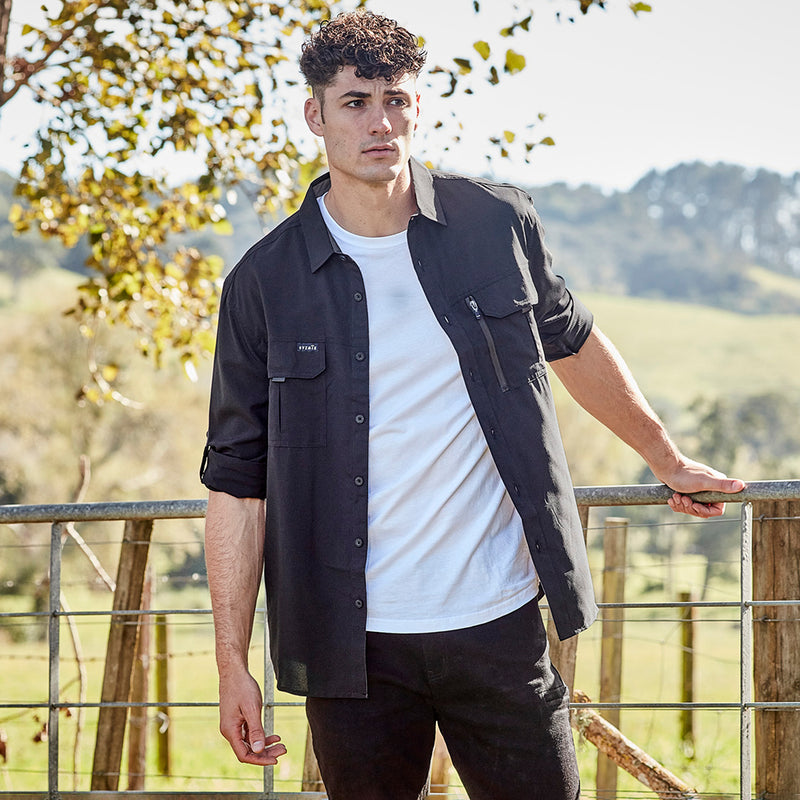 Load image into Gallery viewer, Syzmik Outdoor Long Sleeve Work Shirt, shown worn by a man leaning on a fence, features lightweight polyester fabric, mesh venting, roll-up sleeves, and multiple pockets. Ideal for outdoor work environments.