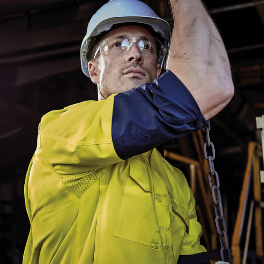 Syzmik Hi Vis Closed Front L/S Work Shirt showcased on a man with a hard hat and safety glasses, highlighting its robust cotton twill fabric and functional design, suitable for demanding work environments.
