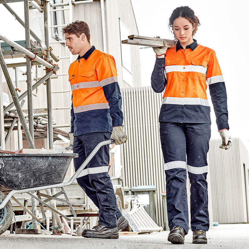 Load image into Gallery viewer, Syzmik Womens Taped Utility Trouser showcased by a man and woman in safety jackets carrying wooden planks, highlighting durable triple-stitched seams, reinforced patches, and reflective tape suitable for construction work.