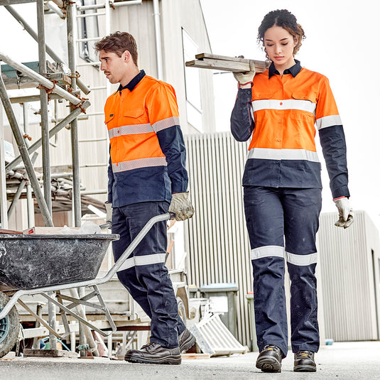 Syzmik Womens Taped Utility Trouser showcased by a man and woman in safety jackets carrying wooden planks, highlighting durable triple-stitched seams, reinforced patches, and reflective tape suitable for construction work.
