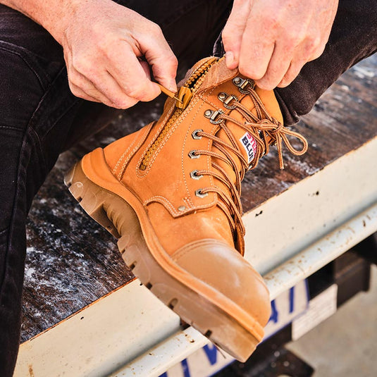 A person zipping up their wheat Steel Blue safety boots. The safety boots have a rugged zip down the side and an outer toe cap for additional safety.