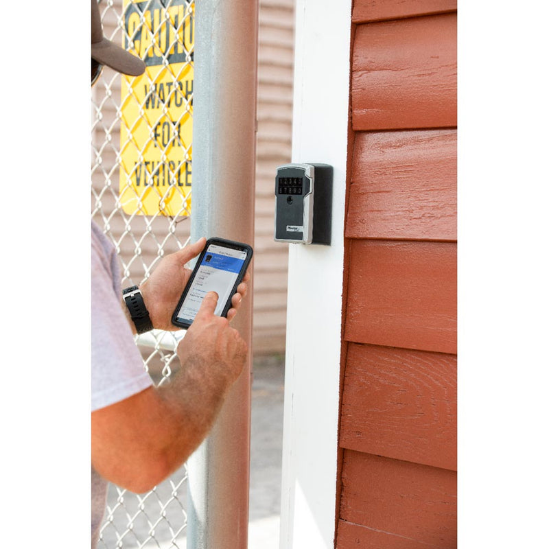 Load image into Gallery viewer, A person uses a smartphone near a Master Lock Bluetooth Wall Mount Lock Box, highlighted by its durable metal body, emphasizing secure, keyless access and monitored entry via Bluetooth technology.
