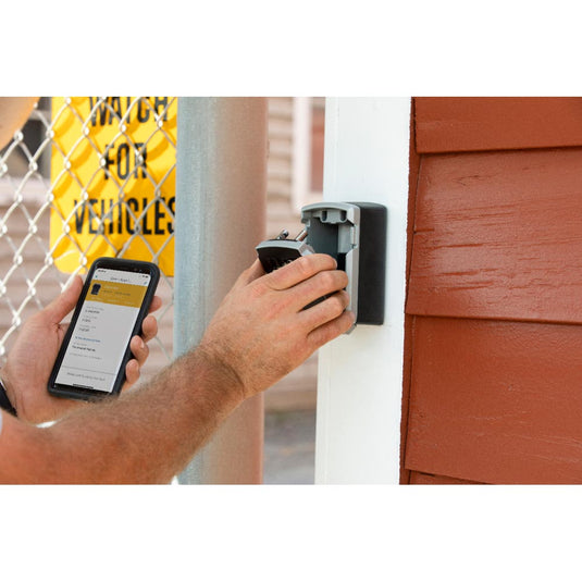 A person uses a smartphone to unlock the Master Lock Bluetooth Wall Mount Lock Box, retrieving a key. The lock box emphasizes security and convenience with Bluetooth technology for managing access.