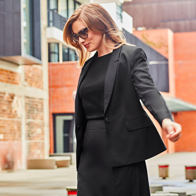 A woman models the Biz Womens Renew Single Button Mid Length Jacket, a sleek, tailored blazer. It's part of a modern suiting range, ideal for versatile, easy-care wear, seen in an urban setting.