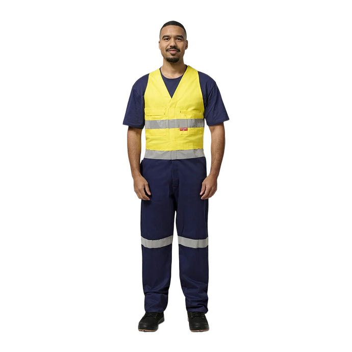 Man models Hard Yakka Easy Action Hi Vis Cotton Bib Overall with reflective strips, navy shirt, and trousers. Features include multiple pockets, elasticized back, ideal for construction safety.