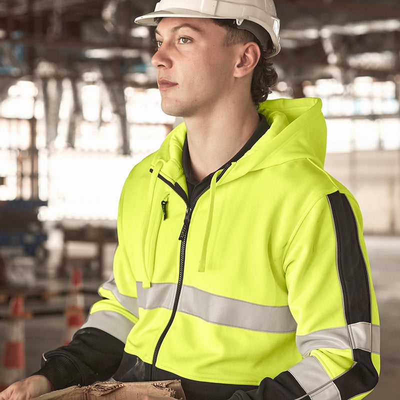 Load image into Gallery viewer, A person wears the Syzmik Unisex Hi Vis Full Zip Stretch Taped Hoodie, featuring reflective stripes, a zip front, and a microfiber-lined hood, standing in an active construction site.
